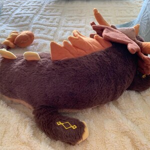 May include: A brown and orange plush toy shaped like a dragon with a long tail and antlers. The toy is lying on a white fluffy surface.
