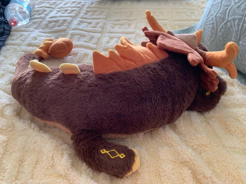 May include: A brown and orange plush toy shaped like a dragon with a long tail and antlers. The toy is lying on a white fluffy surface.