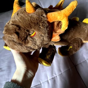 May include: A brown and orange plush dragon with yellow accents. The dragon has a closed eye and a small horn on its head.