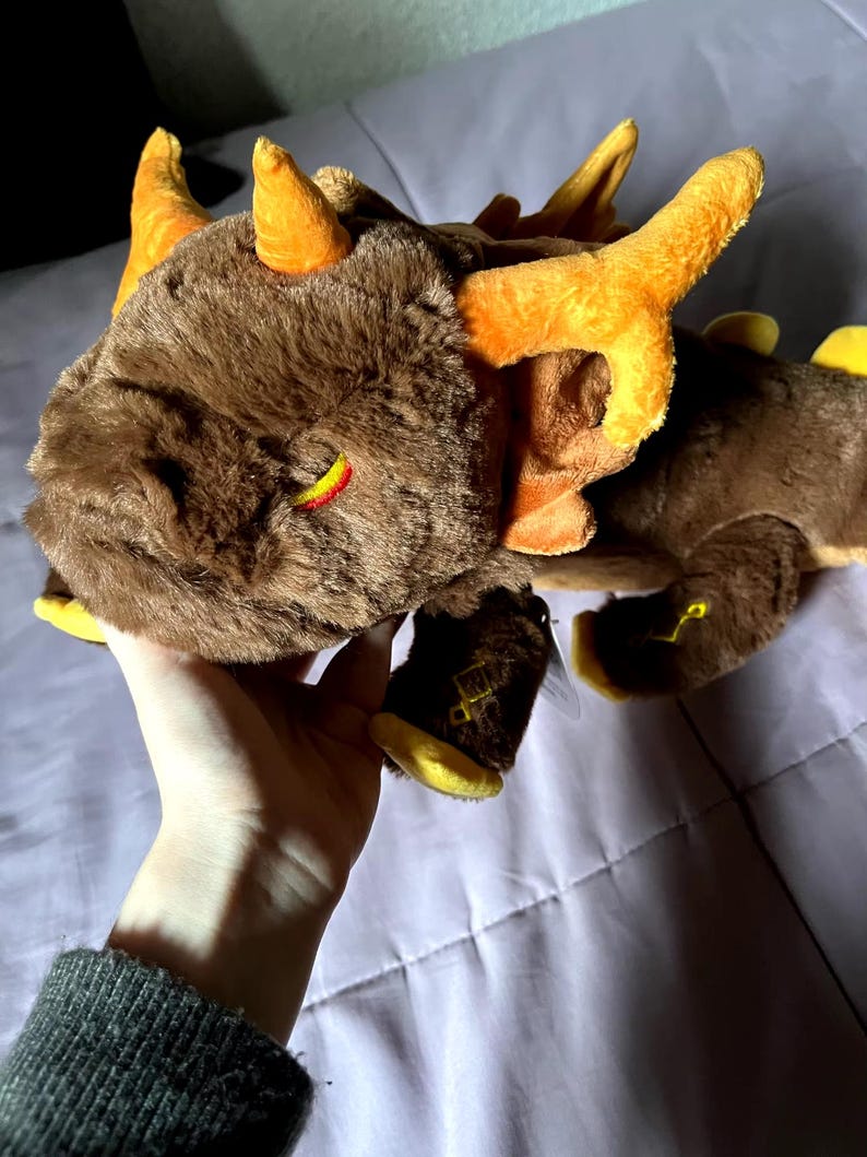 May include: A brown and orange plush dragon with yellow accents. The dragon has a closed eye and a small horn on its head.