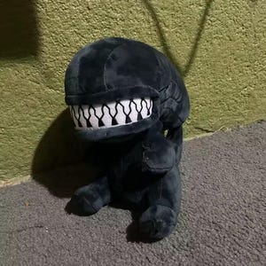 May include: A plush, black Xenomorph toy with a detailed head and a wide mouth filled with white teeth. The toy is sitting on a gray surface, with a green wall in the background. The plush has a tail and four legs.