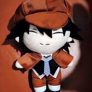 May include: A plush toy of a cartoon character wearing a brown hat, brown jacket, and brown pants. The character has black hair and a white face with a smiling expression.