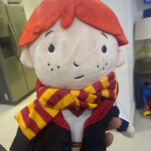 May include: A plush toy of Harry Potter, wearing a red and yellow striped scarf and a black robe. The toy has a red wig and a white face with freckles.
