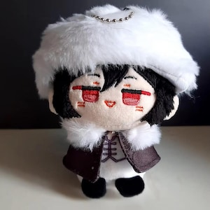 Peut inclure: Un jouet en peluche représentant un personnage aux cheveux noirs, portant un chapeau en fourrure blanche et une veste noire et blanche. Le jouet a des yeux brodés rouges et une bouche souriante.