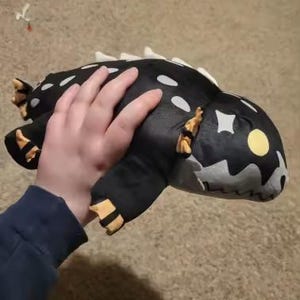 Puede incluir: Un peluche negro con forma de criatura con manchas blancas, aletas dorsales blancas y detalles amarillos en los pies y los lados. El juguete tiene ojos amarillos y una cara gris. El peluche está siendo sostenido.