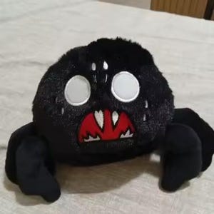 May include: A black plush spider toy with white eyes and a red mouth. The spider has four black legs and a round body. The toy is designed to look like a cartoon spider.