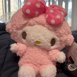 Pode incluir: Um brinquedo de pelúcia com pelo rosa claro, encaracolado e texturizado, com pelo branco nas mãos e nos pés. O personagem apresenta um grande laço rosa brilhante com bolinhas brancas na cabeça, olhos ovais castanhos escuros e um nariz amarelo.