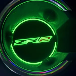 Puede incluir: Un portavasos de coche con iluminación verde brillante, con las letras "ZR2" en blanco y rojo. El portavasos tiene un diseño circular con un contorno verde brillante. El fondo es oscuro.