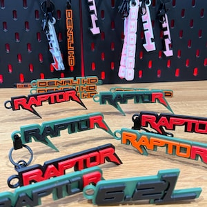 Może przedstawiać: Zbiór kolorowych breloków z napisami "RAPTOR" i "DENALI HD". Breloki są wykonane z miękkiego, elastycznego materiału i mają trójwymiarowy design. Breloki są prezentowane na czarnej tablicy z kołkami.