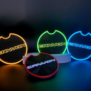 May include: A collection of illuminated car coasters with the word "CANYON" in white, blue, and red. The coasters have a textured surface and a circular shape with a curved cutout. The coasters are lit with orange, green, and blue lights.