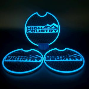 May include: Three circular car cup holder inserts featuring a glowing neon blue border and the text HIGH COUNTRY with a mountain graphic. Each insert has a distinct black textured pattern, including honeycomb, geometric lines, and puzzle pieces.