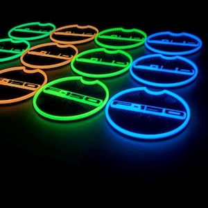 May include: Illuminated F-150 car coasters in green, orange, and blue. The coasters are circular with the F-150 logo. The image is set against a black background, emphasizing the bright colours. The coasters are designed for car cup holders.