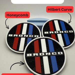 Puede incluir: Tres posavasos circulares con la palabra BRONCO en letras blancas sobre una barra horizontal negra. Los dise&ntilde;os incluyen rayas verticales rojas, blancas y azules sobre patrones de fondo llamados Honeycomb, Hilbert Curve y Octagram Spiral.