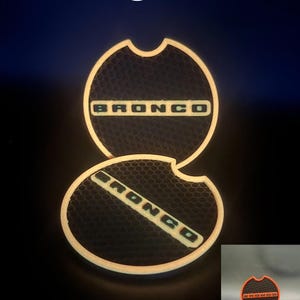 Puede incluir: Dos insertos circulares para portavasos de coche con una textura de panal negra y la palabra BRONCO en letras naranjas. Los bordes de los insertos brillan con una luz naranja, y el texto Orange Glow aparece en la parte superior.