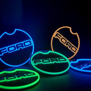Könnte beinhalten: Mehrere beleuchtete Auto-Untersetzer mit dem Ford-Logo in verschiedenen Farben, darunter Blau, Grün und Orange. Die Untersetzer haben eine runde Form mit einer kleinen Kerbe und einer strukturierten Oberfläche.