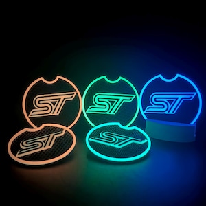 Peut inclure: Sous-verres lumineux pour porte-gobelets de voiture avec les lettres "ST" dans un design stylisé. Les dessous de verre s'illuminent en orange, vert et bleu, créant un effet vibrant sur fond sombre. Les dessous de verre sont ronds avec un dessus incurvé.