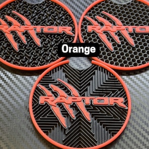 Può includere: Tre sottobicchieri rotondi per auto, neri e rossi, con la parola "RAPTOR" in lettere rosse e un design a forma di artiglio. I sottobicchieri sono su uno sfondo nero in fibra di carbonio.