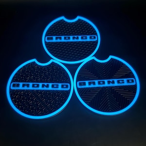 May include: Three black car coasters with a textured surface and a cut-out for easy removal. The word "BRONCO" is illuminated in bright blue on each coaster, creating a striking contrast against the dark background.