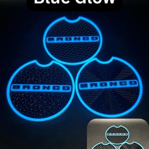 Puede incluir: Tres insertos circulares negros para portavasos de coche con la palabra BRONCO en fuente negrita. Cada inserto tiene un borde brillante de color azul y una superficie texturizada. El texto Blue Glow aparece en la parte superior.