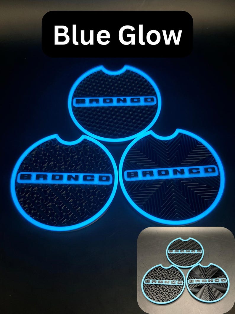 Puede incluir: Tres insertos circulares negros para portavasos de coche con la palabra BRONCO en fuente negrita. Cada inserto tiene un borde brillante de color azul y una superficie texturizada. El texto Blue Glow aparece en la parte superior.