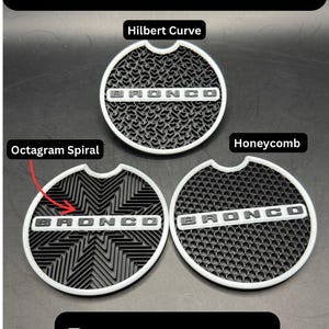 Puede incluir: Tres insertos circulares blancos no luminosos para portavasos de coche con patrones negros. Cada inserto presenta el texto BRONCO en una barra horizontal blanca. Los patrones est&aacute;n etiquetados como Hilbert Curve, Octagram Spiral y Honeycomb.