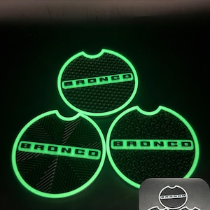 Puede incluir: Tres posavasos circulares negros para portavasos de coche con la palabra BRONCO en una fuente verde brillante. Cada posavasos tiene un patr&oacute;n de superficie texturizado &uacute;nico y un peque&ntilde;o recorte semicircular. Texto Green Glow visible.