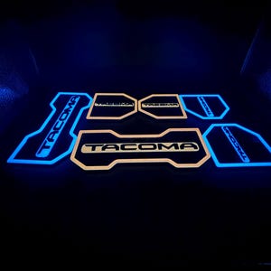 Puede incluir: Alfombrillas iluminadas para coche con la palabra "TACOMA" en un contorno azul brillante. Las alfombrillas son de color beige con la palabra "TACOMA" en el centro. El fondo es negro.