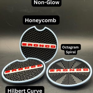 Puede incluir: Tres insertos circulares para portavasos de coche con bordes azul claro y bases negras que presentan patrones distintos etiquetados como Bronco Heritage. Los patrones incluyen Honeycomb, Hilbert Curve y Octagram Spiral. Cada inserto muestra el texto rojo BRONCO.