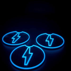 May include: Three glowing blue car coasters with a lightning bolt design. The coasters have a honeycomb pattern and a notch for easy removal. The lightning bolt is outlined in bright blue, creating a striking contrast against the dark background.