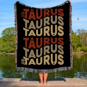 May include: Black woven throw blanket featuring the word "TAURUS" in various colours and sizes. The blanket has a fringed edge and is held up by a person standing on a wooden dock. The background includes a lake and trees.