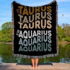 May include: Black woven throw blanket with the words "TAURUS" and "AQUARIUS" in different colours. The blanket has a fringed edge and is held up by a person standing on a wooden dock. The background includes a lake and trees.