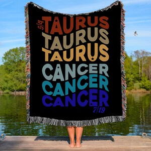 May include: Black woven throw blanket with the words "TAURUS" and "CANCER" in various colours. The blanket has a fringed edge and is held up in front of a person. The background includes a lake, trees, and a blue sky.