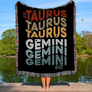 May include: Black woven throw blanket with the words "TAURUS" and "GEMINI" in various colours. The blanket has a fringed edge and is held up in front of a person. The background shows a lake and trees.
