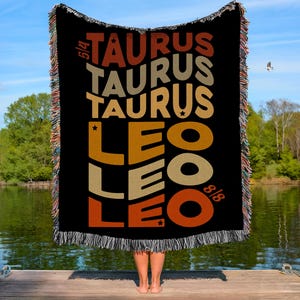 May include: Black woven throw blanket with the words "TAURUS" and "LEO" in a retro font. The blanket has a fringed edge and is held up against a lake and sky backdrop. The dates 5/4 and 8/8 are also visible.
