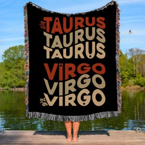 May include: Black woven throw blanket with the words "TAURUS" and "VIRGO" in orange and tan. The blanket is held up by a person on a wooden dock, with a lake and trees in the background.