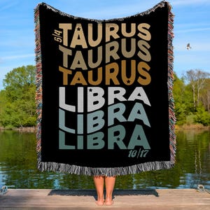 May include: Black woven throw blanket with the words "TAURUS" and "LIBRA" in various colours and fonts. The blanket has a fringed edge and the dates 5/4 and 10/17 are printed on it. A person is holding the blanket on a wooden dock.