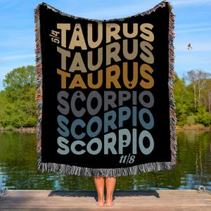 May include: Black throw blanket with the words "TAURUS" and "SCORPIO" in different colours. The blanket has a fringed edge and is held up by a person standing on a wooden dock. The sky and water are visible in the background.