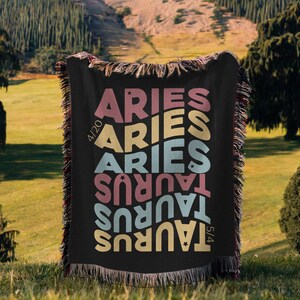 May include: Black woven throw blanket with the words "ARIES" and "TAURUS" in colourful, wavy text. The blanket has a fringed edge and dates 4/20 and 5/4 are printed on the blanket.
