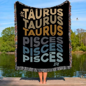 May include: Black throw blanket featuring the words "TAURUS" and "PISCES" in different colours. The fringed blanket is held up in front of a person. The background shows a lake and a blue sky.