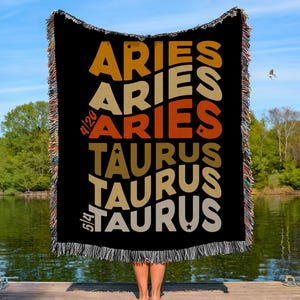 May include: Black woven throw blanket with the words "ARIES" and "TAURUS" in retro lettering. The blanket has a fringed edge and dates 4/20 and 5/14. The background shows a lake and trees.