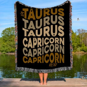 May include: Black throw blanket featuring the words "TAURUS" and "CAPRICORN" in a wavy design. The blanket has a fringed edge with multiple colours. The dates 5/4 and 12/17 are also visible.
