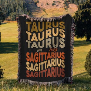 May include: Black throw blanket with the words "TAURUS" and "SAGITTARIUS" in a retro font. The blanket has a fringed edge and the dates 5/4 and 11/23 are printed on it. The blanket is displayed outdoors.