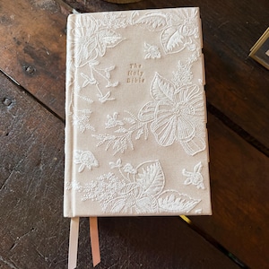 以下が含まれることがあります： 白い花の刺繍が施された、"The Holy Bible"というタイトルのベージュ色の本。表紙には花と葉の細かい模様が施されています。2本のリボンブックマークが見えます。