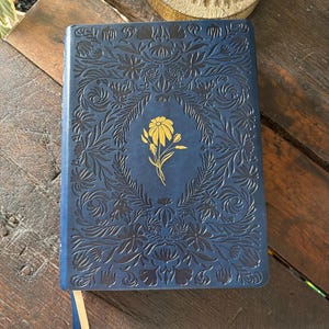 May include: A rectangular navy blue journal with an intricate embossed floral pattern covering the front cover. A single gold-colored flower illustration is centered on the cover. Two thin ribbon bookmarks, one blue and one cream, extend from the bottom.
