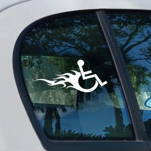 以下が含まれることがあります： 車の窓に貼られた白いビニール製のデカール。車椅子を模したシンボルマークに炎のデザインが組み合わされており、後ろにたなびく炎が動きとスピード感を演出している、アクセシビリティをテーマにしたグラフィックステッカー。