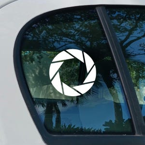 May include: A white vinyl decal featuring a camera aperture design with six distinct blades arranged in a circular pattern. The sticker is applied to the exterior of a car window, displaying a clean, high-contrast graphic against the glass.