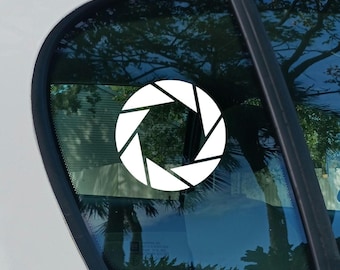 May include: A white vinyl decal featuring a camera aperture design with six distinct blades arranged in a circular pattern. The sticker is applied to the exterior of a car window, displaying a clean, high-contrast graphic against the glass.