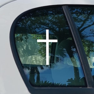 May include: A white cross decal is affixed to the rear window of a vehicle. The window reflects a view of trees and a building, with the cross prominently displayed against the blue-tinted glass.