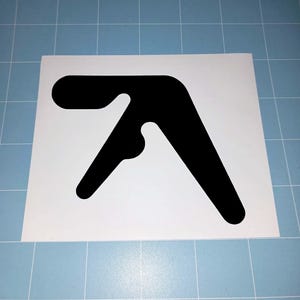 May include: A black sticker featuring the Aphex Twin logo, a stylized, abstract design. The logo is a solid black shape on a white background. The sticker is rectangular and placed on a blue grid surface.