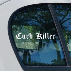 May include: White vinyl decal with the words "Curb Killer" in a gothic font, applied to a car window. The window reflects trees and sky.